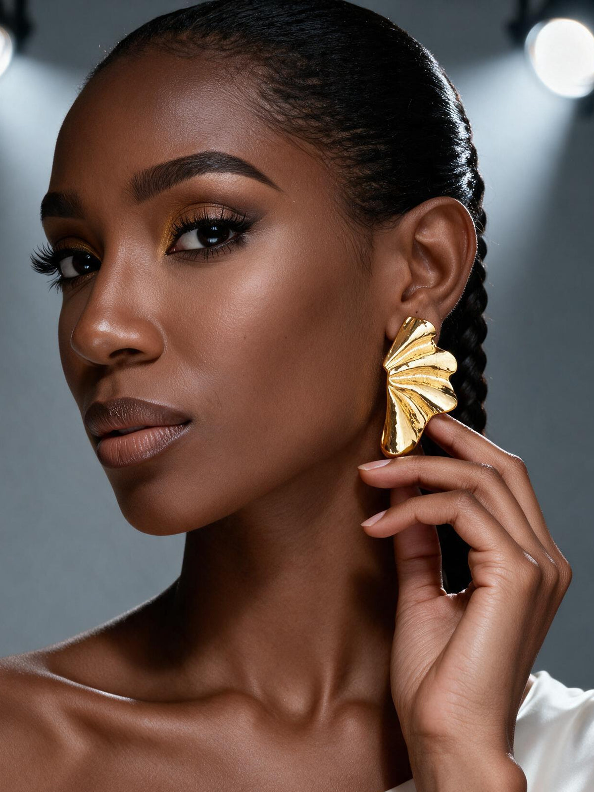 Side profile of a model wearing oversized gold botanical statement earrings inspired by Ginkgo leaves, highlighting the modern sculptural petal silhouette with her hand touching the earrings.