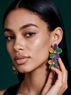 Close-up of oversized floral petal earrings featuring purple glitter, emerald green enamel, and gold accents worn by model.