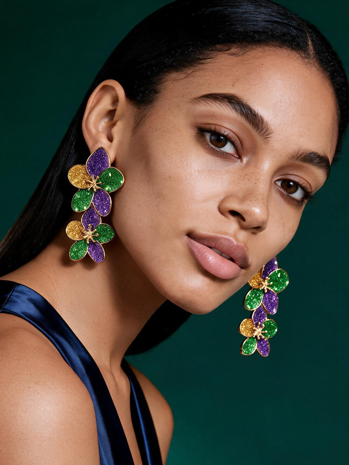 Model wearing colorful Mardi Gras-inspired flower statement earrings in purple, green, and gold with elegant green satin dress.