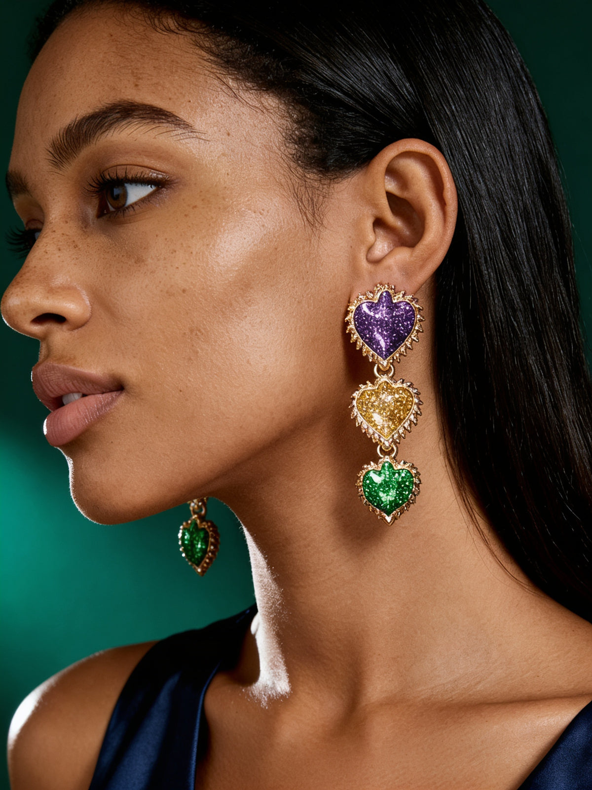 Model wearing colorful Mardi Gras-inspired flower statement earrings in purple, green, and gold with elegant green satin dress.